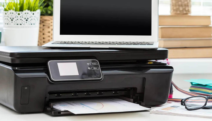 Impressora multifuncional preta em mesa de escritório com notebook, papéis e óculos ao lado